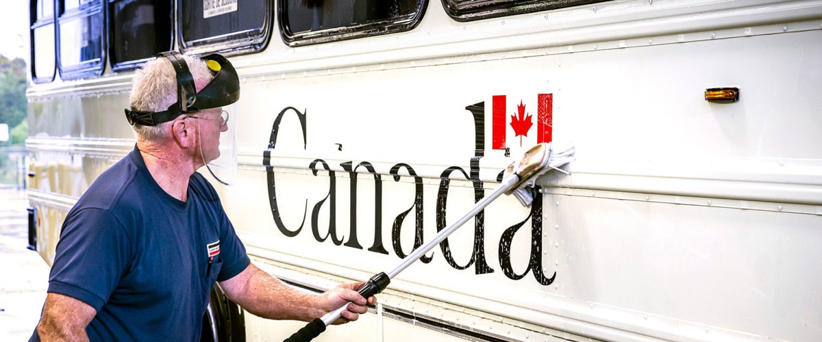 CBO employee with a faceshield washing a large Government of Canada bus with a nozzle and brush.
