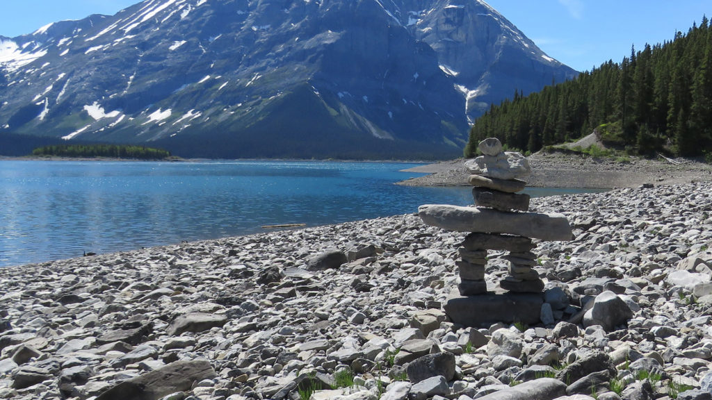 An inuksuk on a bed of rocks beside a lake at the foot of a mountain.